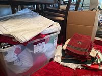 Bin with various Christmas towels, tablecloths, and placemats stacked next to cardboard boxes on red carpet floor
