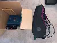Photo showing a box with board games including Trivial Pursuit Master Game and tennis rackets in a carrying bag alongside a pool cue.