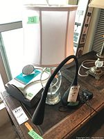 Photo shows a black flexible gooseneck desk lamp, a crystal table lamp with a rectangular white fabric shade, and another black adjustable desk lamp, arranged on a wooden surface near a window.
