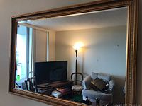 Full view of the rectangular mirror mounted on a wall reflecting room furniture and decor including TV, chairs, and a standing lamp.