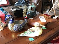 Photo showing three decorative duck figures: Murano glass duck with brass overlay, signed wooden decoy duck, and pottery duck with blue scale details.