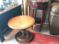 View of both round wood veneer side tables, showing the pedestal table in front and the taller table with drawer and shelf behind.