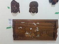 Photo showing two Asian wood carved masks mounted on wall above a wooden tray with mother of pearl inlay depicting a naturalistic scene with figures and a tree.