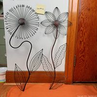 Two large metal wireframe flower sculptures with leaves and stems, bronze finish, standing against a wall with hardwood trim.
