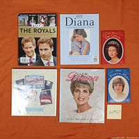 Top view of six books on the British Royal Family arranged on an orange surface. Includes three books about Diana, one on Queen Elizabeth II, the Royal Baby Book, and a 'People' magazine titled 'The Royals'.