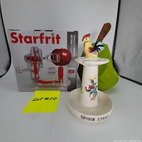 Starfrit apple peeler in original box, vintage ceramic rooster spoon stand holding a wooden spoon, and carved wooden pear shown together on white background.