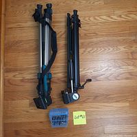 Two tripods, one Optex and one Focal, laid on a wooden floor with extra tripod parts in a blue plastic container.