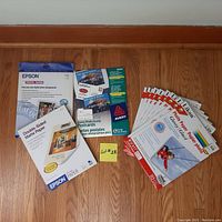 Photo showing a lot of inkjet photo paper packages arranged on the floor against a white wall. Includes Epson Double-Sided Matte Paper, Avery Glossy Photo Quality Postcards, and multiple LOKON Photo Paper packages.