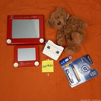 Photo showing two red Etch A Sketch toys (regular and travel size), a white Bicentennial View-Master viewer, a brown teddy bear, a new Hot Wheels Drop Stars die cast model in packaging, and a metal Ford mini gas pump model on an orange surface.