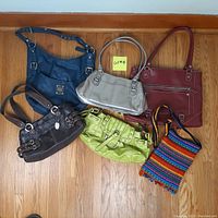 Photo showing six handbags and purses arranged on a wooden floor. The bags vary in color and style.