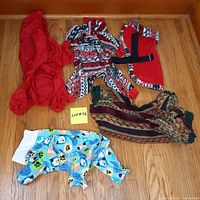 Five small dog clothing items: red raincoat, multi-patterned fleece onesie, red fleece vest, patterned pullover, and cartoon print onesie on wood floor.