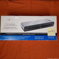 Photo showing the laminator's original box with product image and description stating it is a 13 inch laminator with hot and cold settings, overheat protection, and accepts pouch thickness up to 80-125 microns.