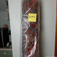 Side view of one rolled Alexa area rug in original plastic packaging with a 'Lot #2' yellow label attached