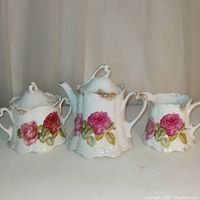 Front view of three-piece porcelain set including teapot in center, sugar bowl with lid on left, and creamer on right. All pieces decorated with red roses and embossed Art Nouveau style shapes.