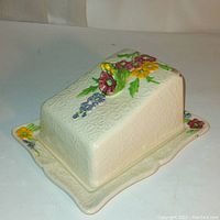 Overall front angled view of the rectangular ceramic cheese dish and lid, showing embossed heart patterns and colorful floral designs on the lid and base edges.