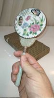 Hand mirror held in hand showing round porcelain back with colorful butterfly and flower design and jade handle.
