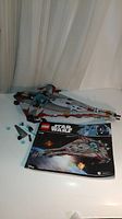 Assembled Lego Arrowhead ship with smaller ship piece and instruction manual shown on white surface against curtain background.