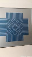 Close-up of the blue geometric abstract optical art print on gray background showing part of the design and silver frame edge.