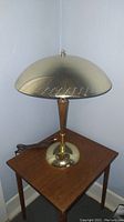 Full view of the vintage atomic style table lamp with domed brass shade and teak wood stem on a small wooden table.