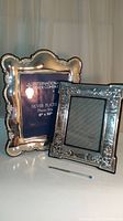 Pair of ornate photo frames, one silverplate with scalloped edges, and one sterling pewter with floral embossing on white surface.