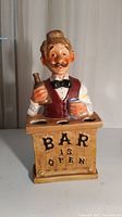 Front view of the vintage ceramic bar man assistant holder holding a bottle and a glass, with text 'BAR IS OPEN' on the base.