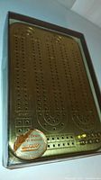 Top view of the solid brass cribbage board in original box showing the full playing surface with peg holes and start area, label indicating "Solid Brass" and "Die Cast."