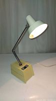 Photo showing the vintage Mobilite desk lamp with off-white metal shade, cream base, chrome adjustable arm, lamp powered on with warm light.