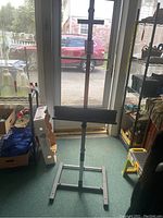 Full view of the metal artist easel standing upright near a glass door, showing the entire height of the easel including the height adjustment and base support structure.