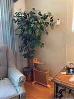 Photo showing a faux leafy tree about 57 inches tall and approximately 37 inches wide standing in a woven basket planter, alongside a wooden magazine rack roughly 15 inches wide. Positioned next to an armchair and a wooden side table with a lamp.