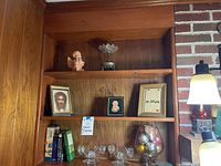 Wooden shelving unit with candle sticks, religious framed pictures, books, and a bowl of Christmas ornaments arranged on three shelves.