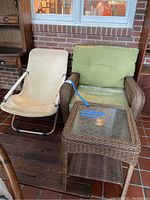 Rattan outdoor armchair with green cushions next to matching rattan side table with glass top and beige canvas folding chair.