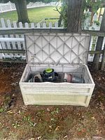 Outdoor white diamond-pattern plastic storage box with lid open showing multiple planting pots inside