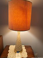 Lit lamp showing tall tapered base with cream color and linear mid-century pattern, tan shade casting warm light.