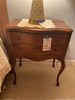 Nightstand with a lamp and lace doily on top, showing curved drawer fronts and wooden knobs.