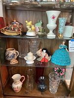 Three shelves containing an assortment of vases, porcelain dolls, decorative figurines including an Asian boat and Japanese doll, and two cream-colored Lenox vases.
