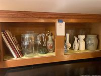 Wooden shelving with Pyrex glass pour-through coffee pot, ceramic vases including floral-painted and plain white, and a ceramic jar labeled 'Slate Pottery'. Also includes small green flower frog device.