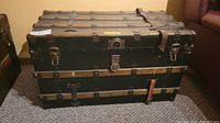 Front view of antique steamer trunk showing black metal exterior with wood slats, metal clasps and leather handles.