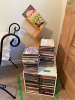 Stacked music CDs on and inside a white laminate cube storage unit with a wood wall-mounted CD rack leaning nearby.