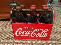 Front view of the metal Coca-Cola carrier filled with six vintage glass bottles with red caps sitting on carpet.