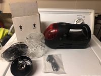 Handheld air compressor (black with red translucent section), unpackaged wall lamp glass cover, lamp mounting base, metal hooks in plastic, and original box on white table
