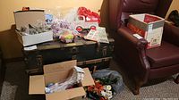 Wide view showing boxes of various craft and holiday items including stuffed plastic eggs, floral scrapbooking pieces, and stuffed bags on a wooden trunk and floor near a chair.