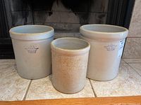 All three stoneware crocks displayed on tile floor in front of fireplace showing variety in size and condition