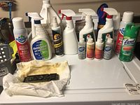 Photo showing assorted household chemical spray bottles and aerosol cans arranged on a white surface with a cloth and small black object.