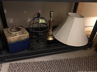 Photo showing ceramic lamp base with beige and blue lava-style finish, black metal cage tea light holder, wood and metal lamp base, and white fabric lamp shade on black shelf.