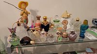 Wide view of 16 assorted vintage and decorative glass perfume bottles displayed on a shelf, including Avon and jeweled themed bottles plus brass boot and silverplate peacock.