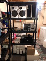 Front view of black resin utility shelving unit filled with various objects (not included) showing five shelves and tubular corner supports.