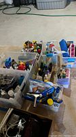 Photo of multiple clear storage bins filled with assorted bottles, sprays, and household products arranged on basement floor.