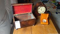 Wide view showing all three items on table: wooden mantle clock, travel clock in orange case, and open wooden trinket box with red fabric inside.