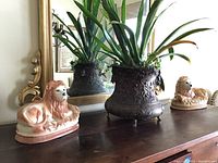 Copper pot containing a green live plant, flanked by two ceramic lions sitting on a wooden surface in front of a mirror.