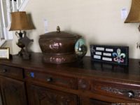 Wide shot showing large lidded copper tub placed on a wooden sideboard with lamps on either side, hand-painted large wooden egg beside the tub, and framed decor plaque.